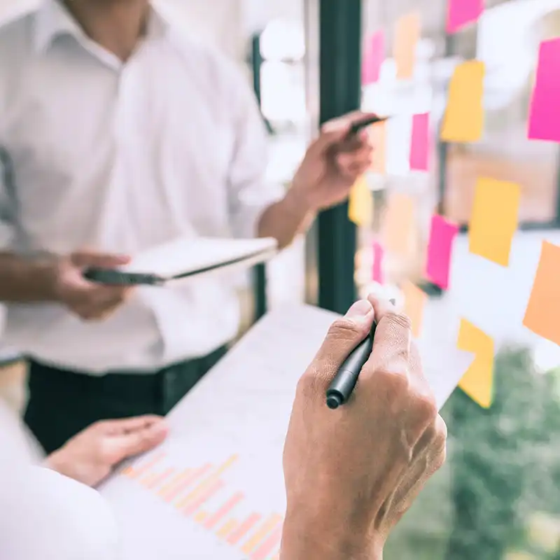 Two people brainstorming with colorful sticky notes on a glass wall, representing Artmedia Digital's collaborative approach in market research.