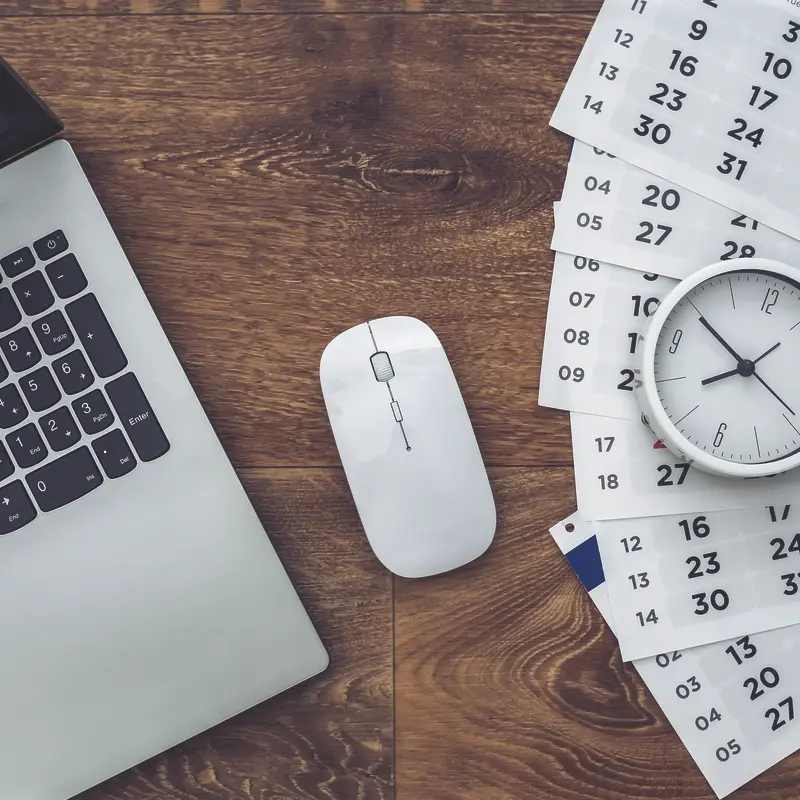 Laptop and mouse on a wooden desk beside scattered calendar pages and a clock, symbolizing Artmedia Digital's time-efficient web design services.